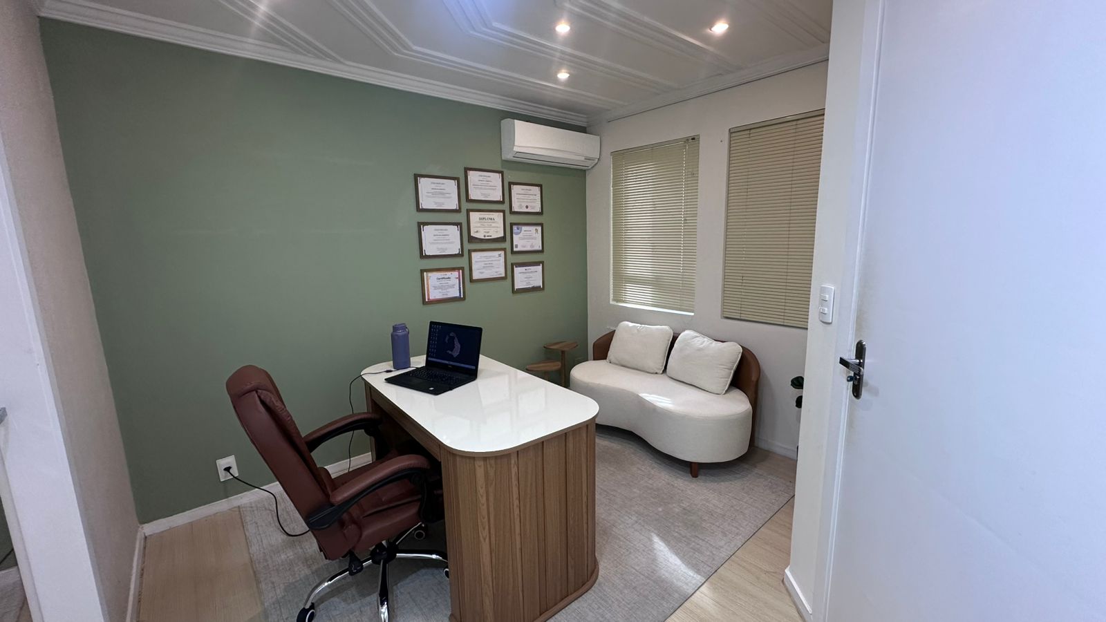 Sala de atendimento — mesa, poltrona e diplomas na parede verde-sálvia